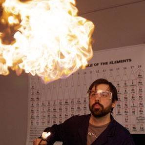 Eric Camp wearing a lab coat and safety googles watching a beach ball sized flame above his head during a lab demonstration in 2016.