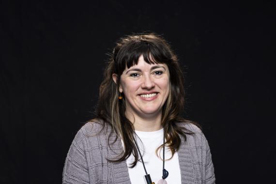 Colleen Craig pictured smiling, in front of a black backdrop, with long dark hair past her shoulders and bangs, wearing a grey cardigan, white top, and long necklace with chunky beads.
