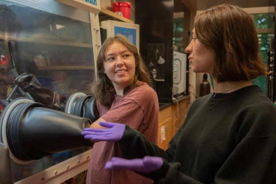 A scientist uses a glove box while another scientist looks on.