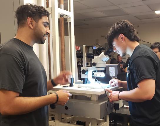 Arturo Lopez peers through a microscope as Zeeshawn Kazi stands to the left.