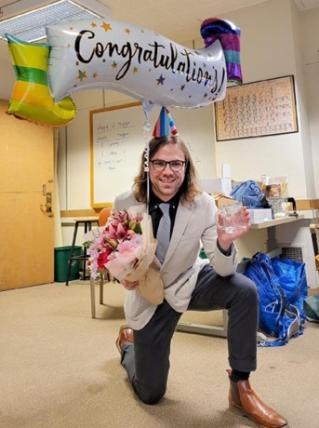 Tim holds up a balloon saying “congratulations”, a bouquet of flowers, and a mug with “Dr. Trinklein” written on it while wearing a party hat.