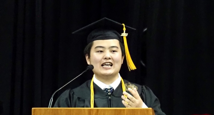 William Wu wearing a graduation cap and gown standing behind a lectern