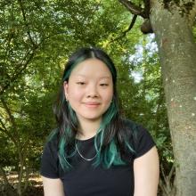 Mary Deng in a black tshirt near a tree smiles with a closed mouth