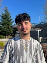 Omeed Yazdani wears a short sleeved shirt, gold chain, and goatee, pictured outside on a day with blue skies