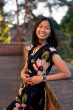 Jolene Nguyen wears a floral dress and leans against a half wall with trees in the background