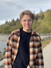 Benjamin Mustonen wears a flannel shirt over a black tshirt and is pictured at a river bank or lake shore