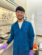 Adrian Guerrero poses in a chemistry lab wearing googles, a blue lab coat, and purple nitrile gloves.