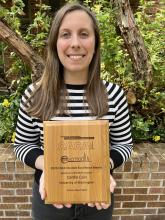Caitlin Cain holds a wooden plaque for her CACA Student Excellence Award. She is wearing a sweater with black and white horizontal stripes and black jeans. She is standing outside in front of a short brick wall and shrubbery.