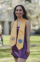 Elouisa Flores wears a purple dress and a gold stole, standing in the Quad with cherry blossom trees in the background