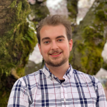 Jacob Russell wears a plaid collared shirt and a short beard. Jacob is pictured near a cherry blossom tree.