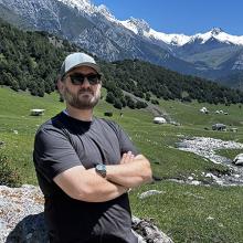 Johnathan Cox with arms folded, with snowy peaked mountains of Kyrgyzstan behind him.