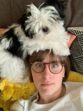 Katie Nowlin lounges on the couch with a small fluffy white and black dog resting on her head.