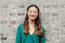 Vivian Li in a green blouse with white polka dots, smiling and standing in front of a gray brick wall