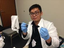 Zicong Li in a lab coat, wearing blue disposable gloves, holding a sample