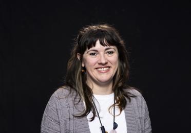 Colleen Craig pictured smiling, in front of a black backdrop, with long dark hair past her shoulders and bangs, wearing a grey cardigan, white top, and long necklace with chunky beads.