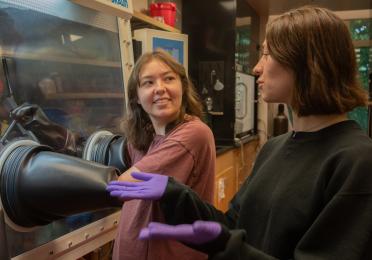 A scientist uses a glove box while another scientist looks on.