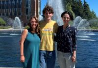 Three people stand in front of a large fountain on a sunny day
