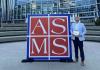 Nick holds a plaque while standing next to a large sign that say ASMS (for the American Society for Mass Spectrometry)