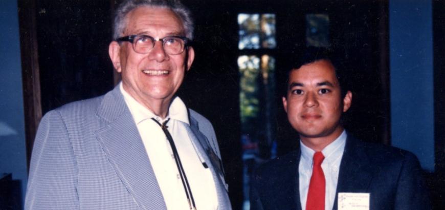 Lingafelter, an older white man wearing glasses and a bolo tie, stands to the left of Santarsiero, a young Latino man wearing a dark suit and red tie.