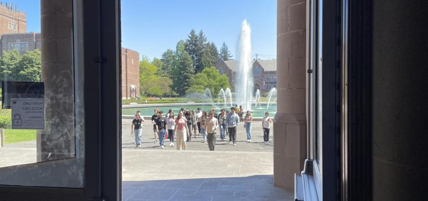 A group of people enter Bagley Hall. Drumheller Fountain is in the background.