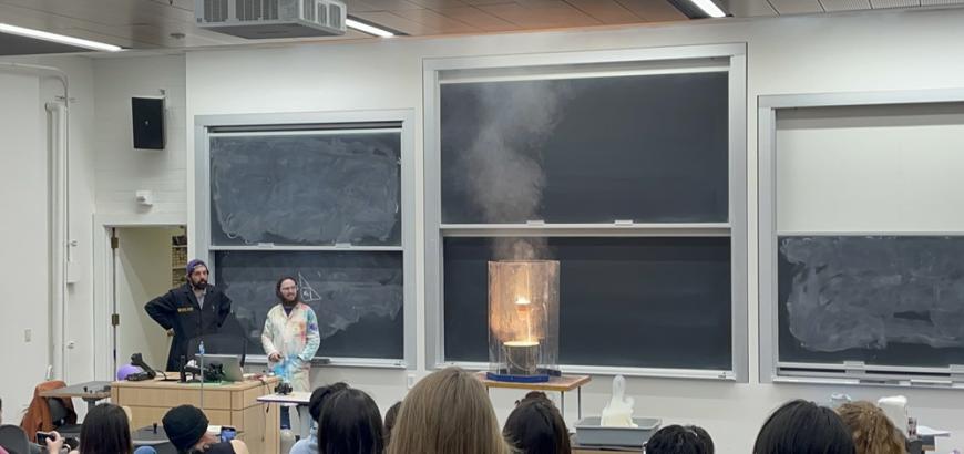 Two people in lab coats stand aside an exothermic reaction demonstration as the audience watches.