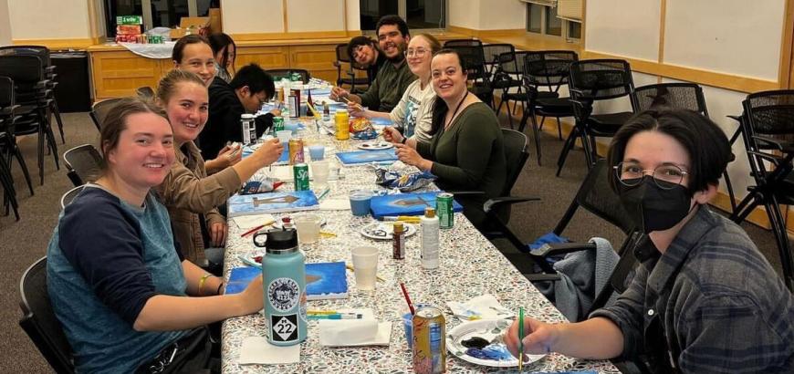10 people sit at a long table painting on canvases.